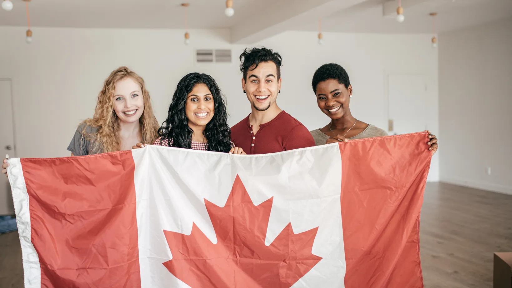 Four smiling friends holding a large Canadian flag indoors