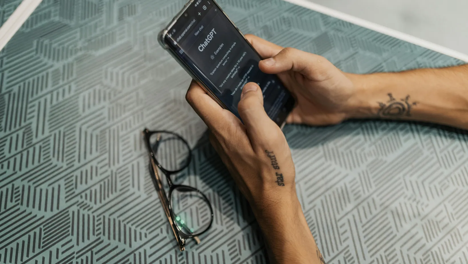 Hands holding a phone open to a new ChatGPT chat, with glasses on the table