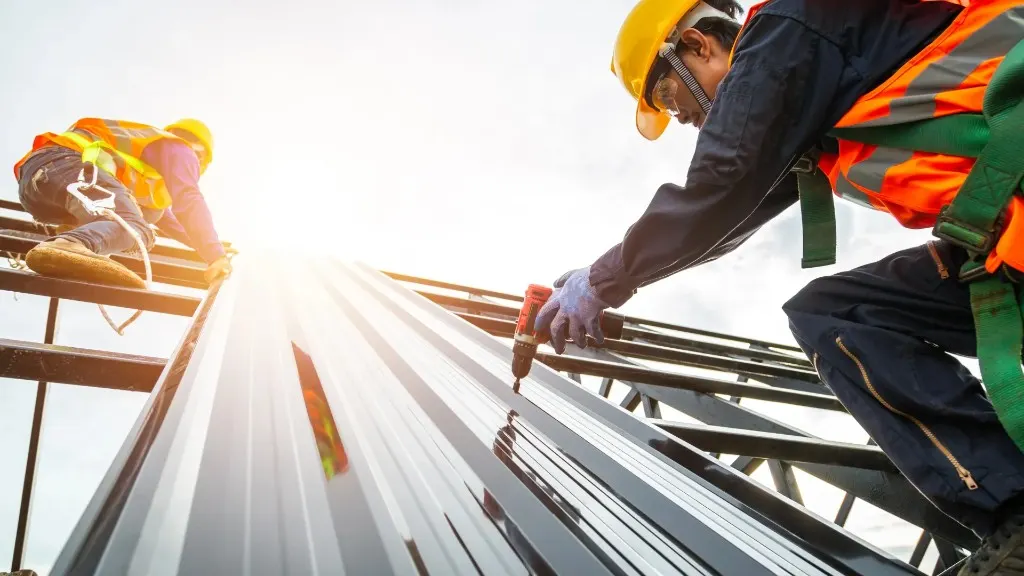 Two contractors in safety gear fastening corrugated metal cladding panels on a roof frame