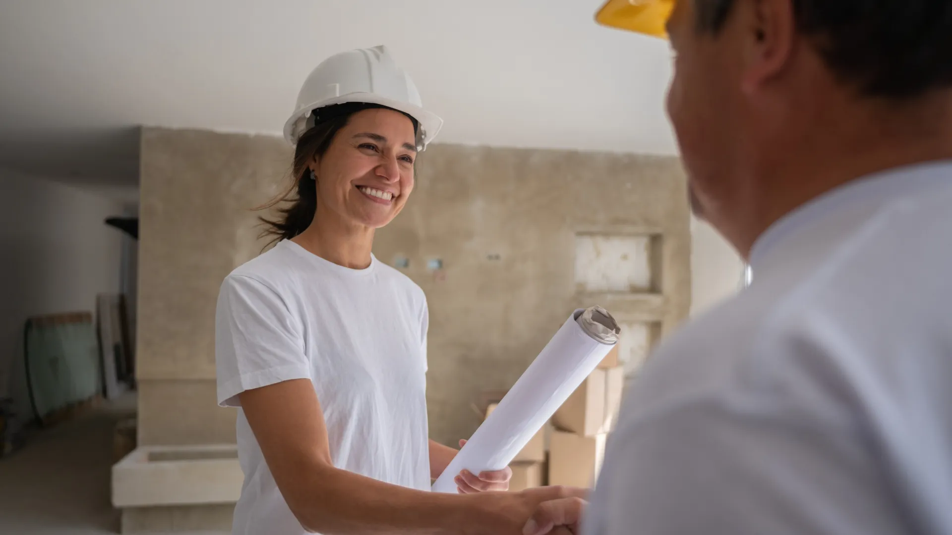 A contractor in a white hard hat holds rolled plans and shakes hands on a renovation site