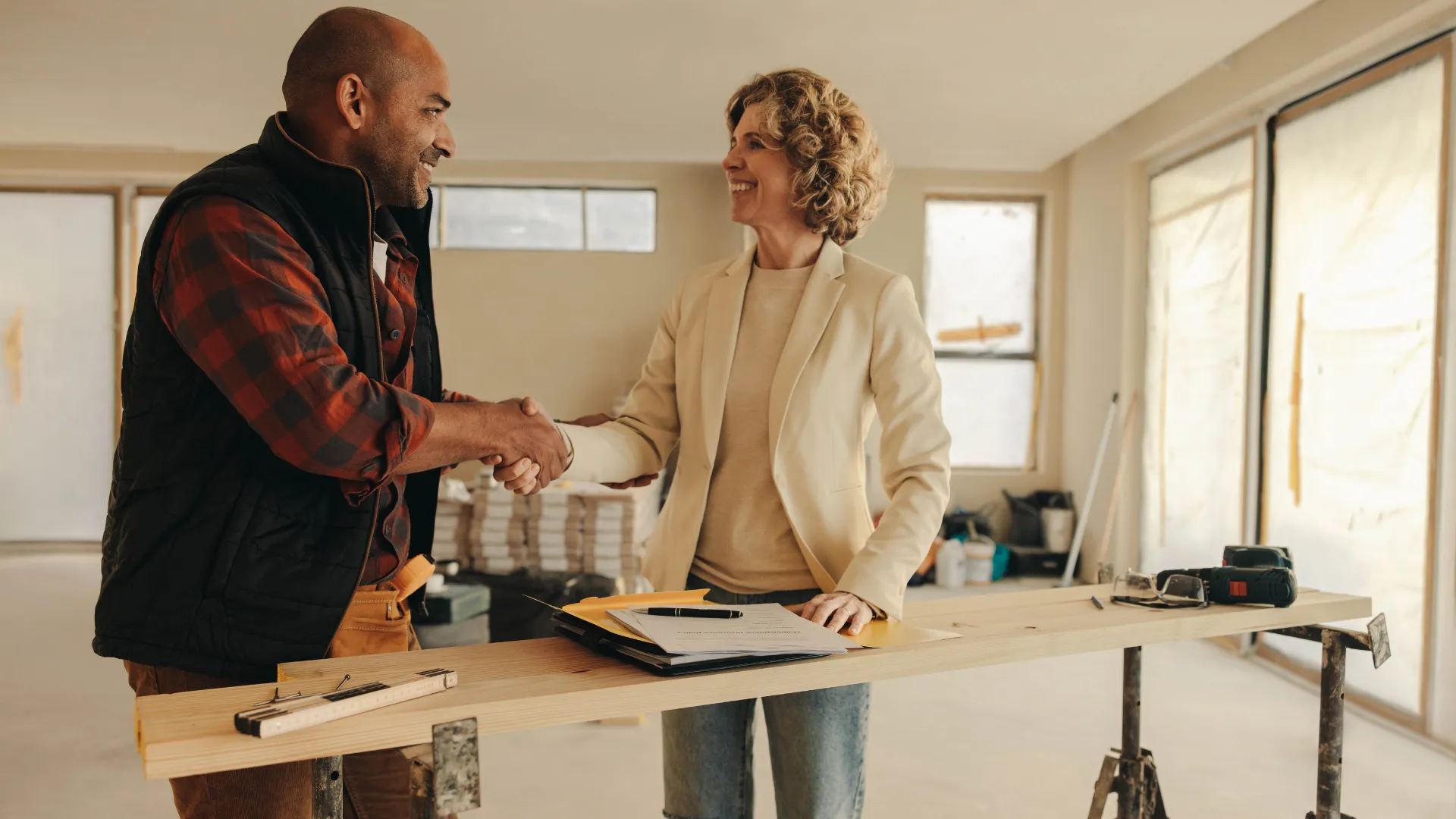 A contractor and a client shake hands over plans inside a renovation