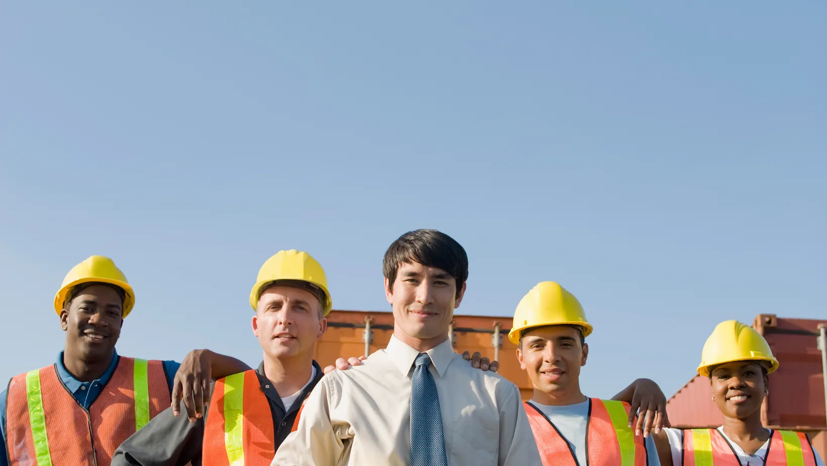 Construction crew in yellow hard hats and safety vests standing with a manager in a shirt and tie