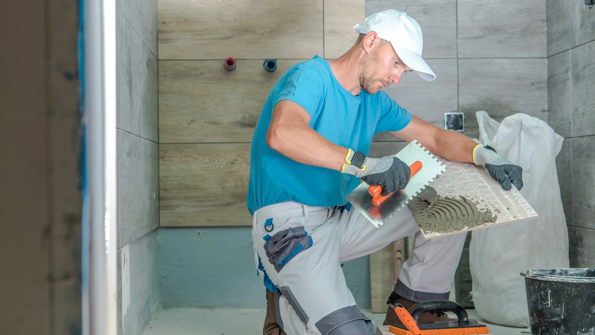 A tiler in a white cap spreads mortar on a large tile
