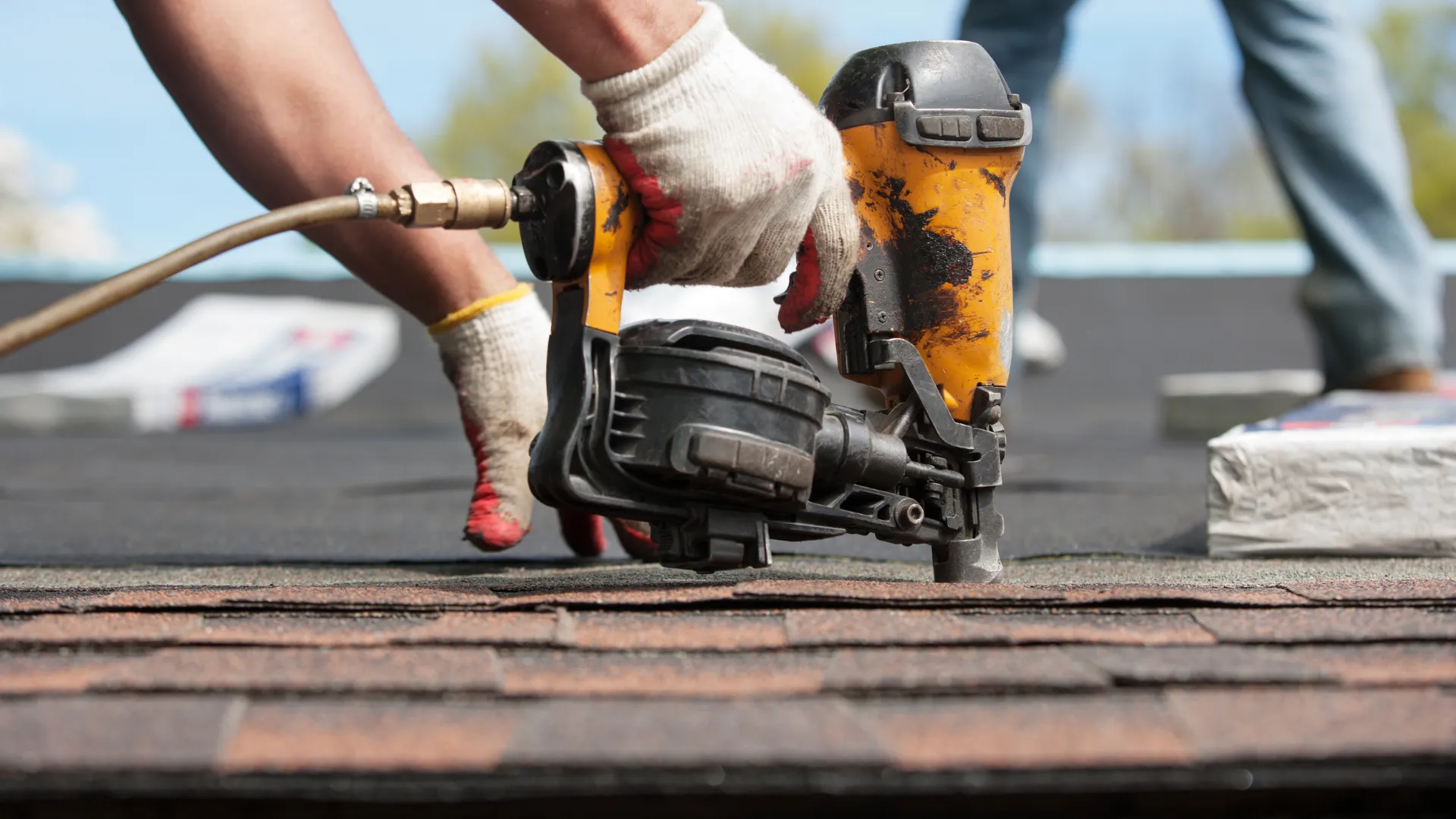 A gloved hand drives a nail gun into roof shingles