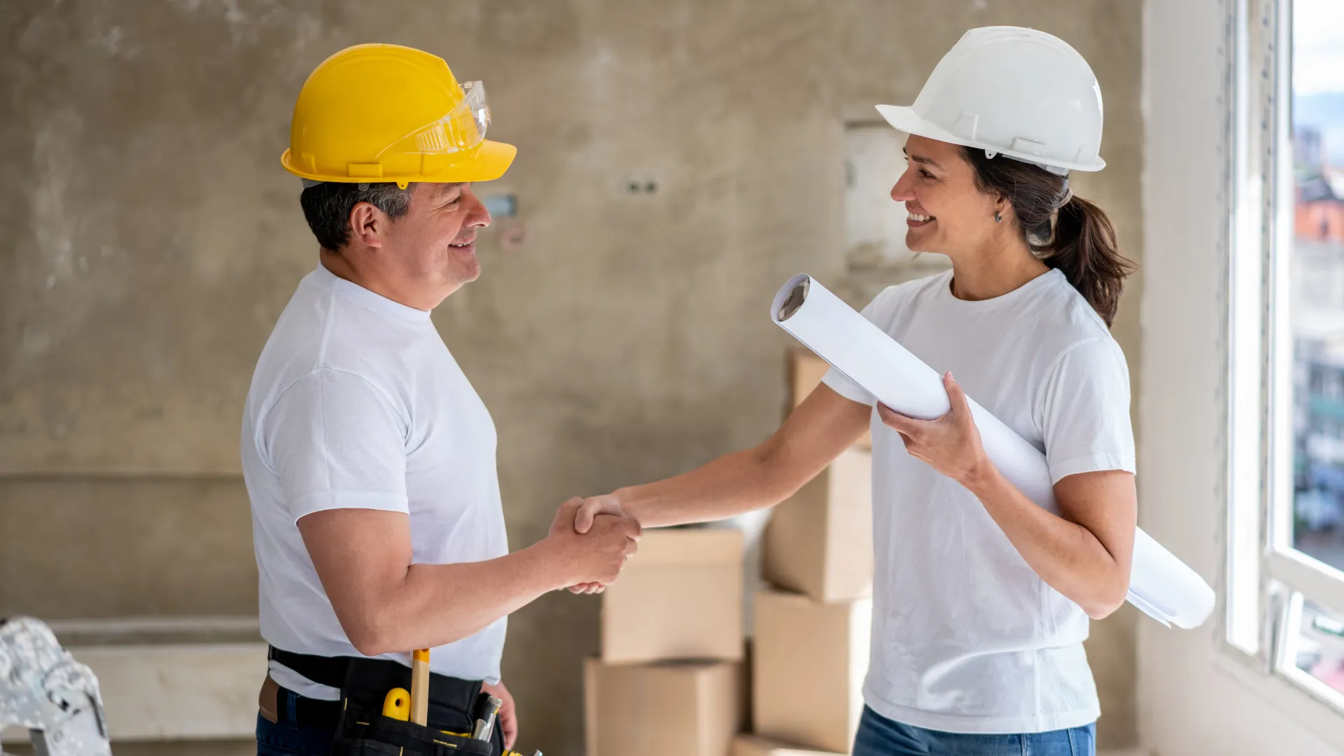 A contractor in a yellow hard hat shakes hands with a colleague holding blueprints