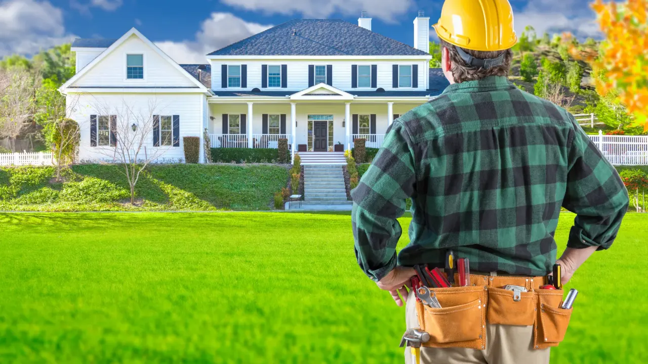 Contractor in a hard hat and tool belt looking toward a two-story colonial home in a suburban neighborhood