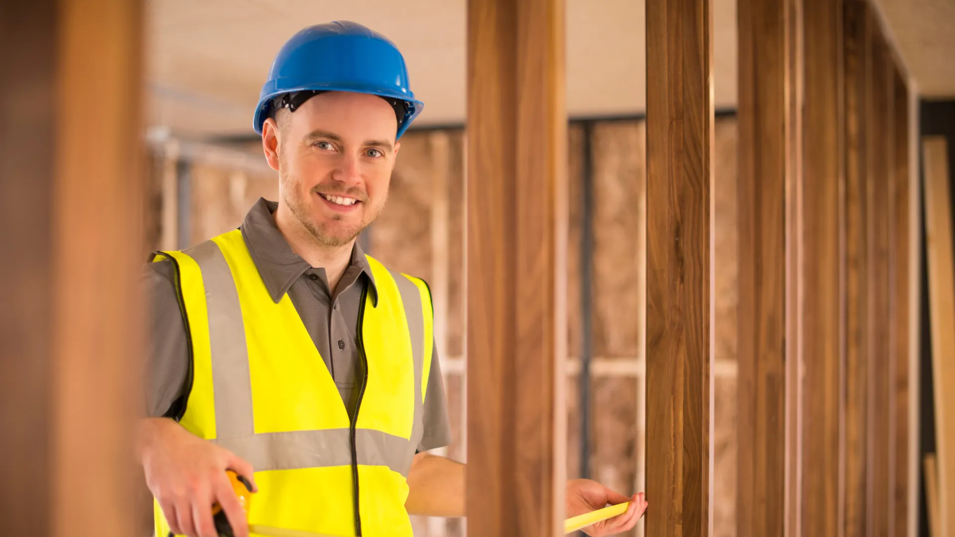 Contractor in a blue hard hat and safety vest measuring timber wall framing