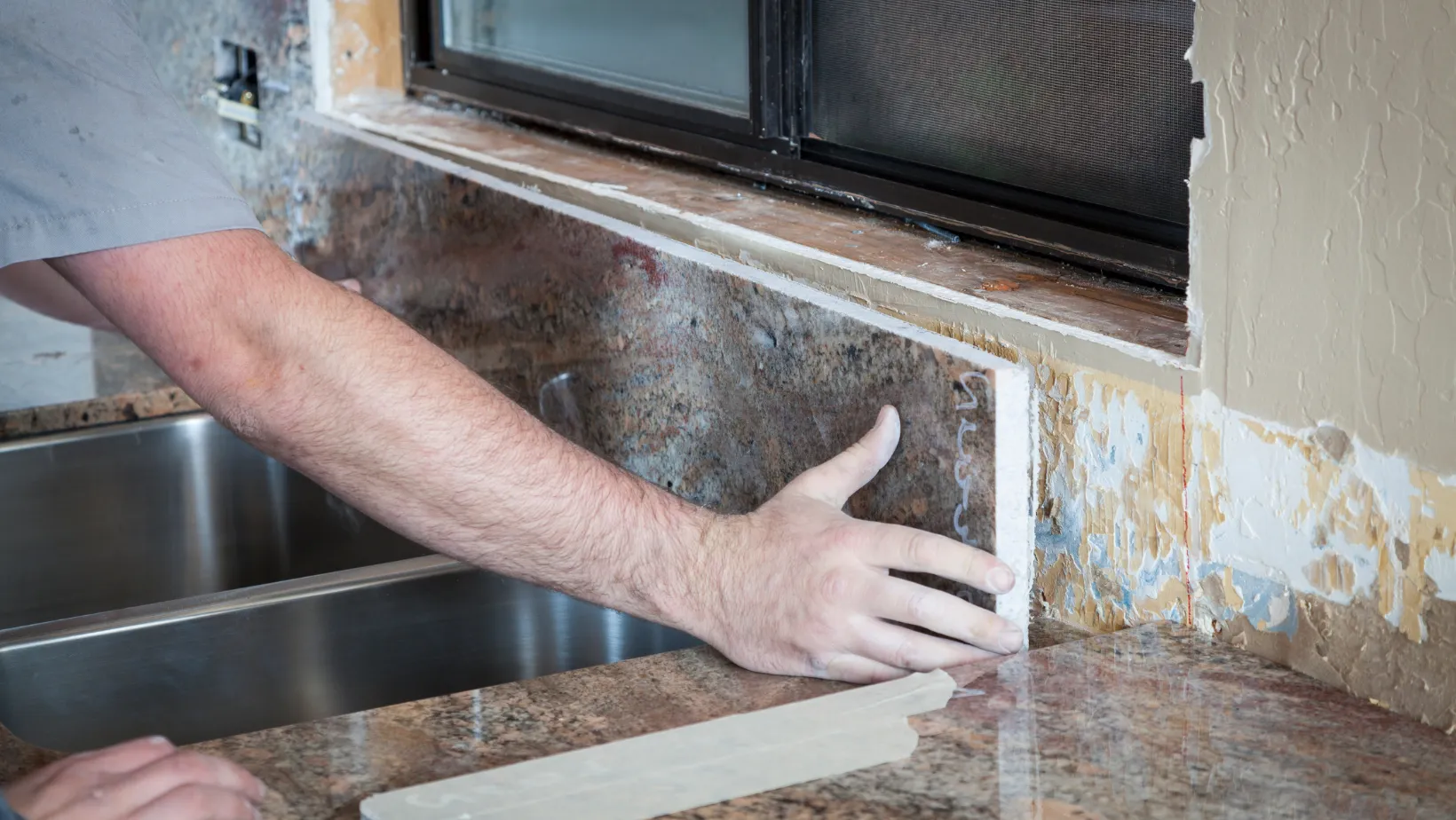 A hand fitting a granite backsplash piece beside a kitchen sink.