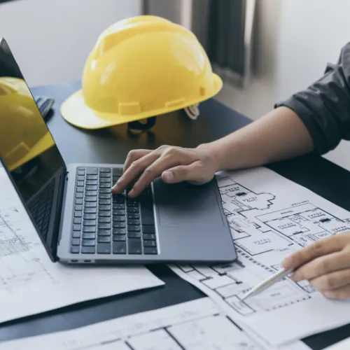 Contractor reviewing floor plans on a laptop beside a hard hat