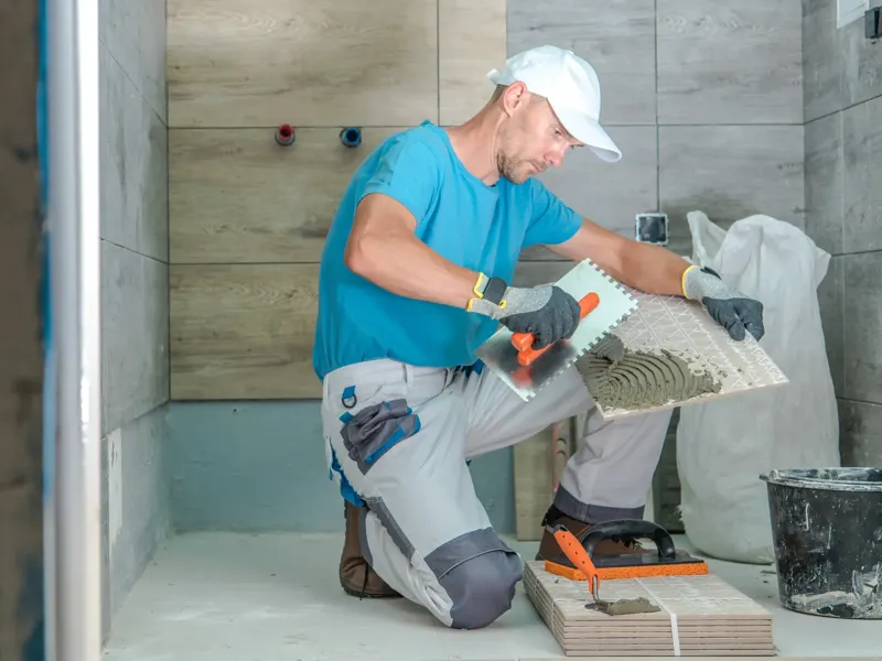 A bathroom remodeler spreading cement on the back of a tile in a bathroom.