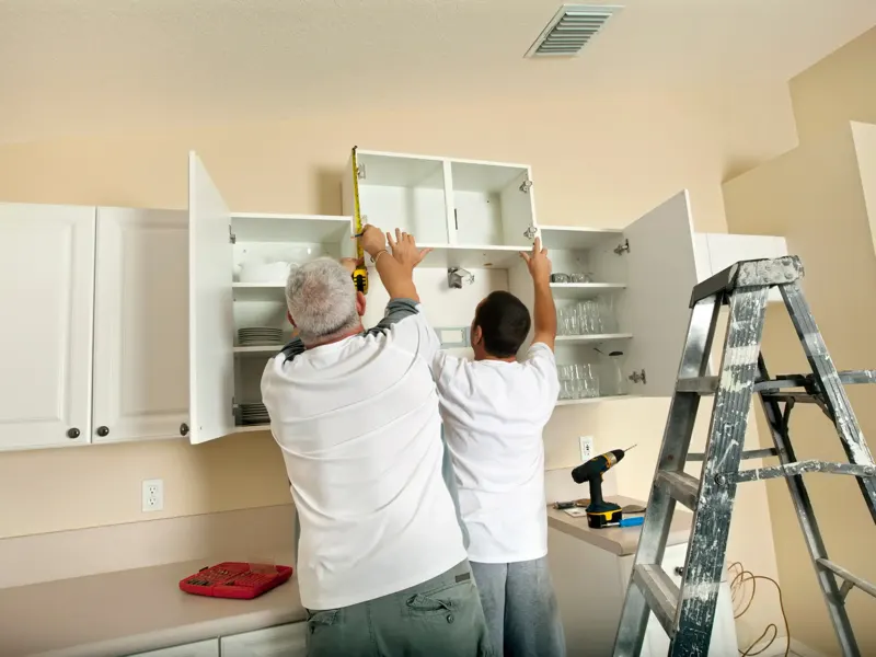 Two kitchen remodeling contractors measuring and installing a new kitchen cabinet on a kitchen wall.