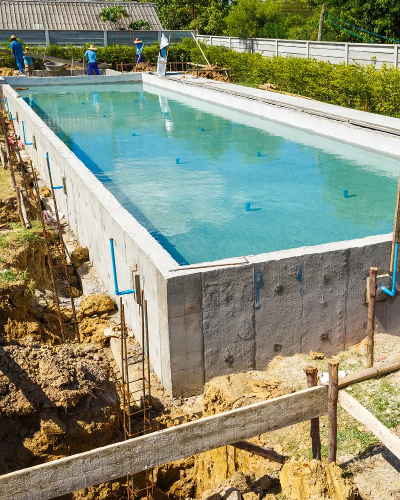 In-ground concrete pool shell filled with water during construction, with plumbing pipes and crew working on site
