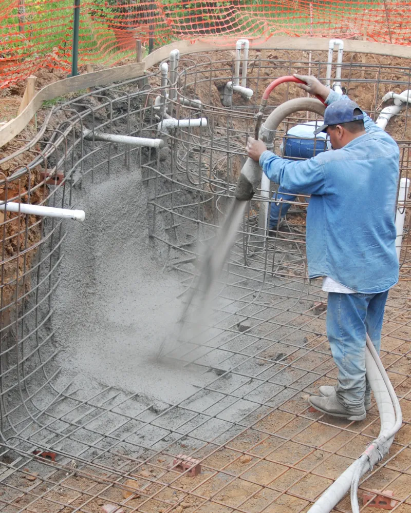 Pool construction crew spraying shotcrete onto rebar for a custom inground pool shell at an active job site