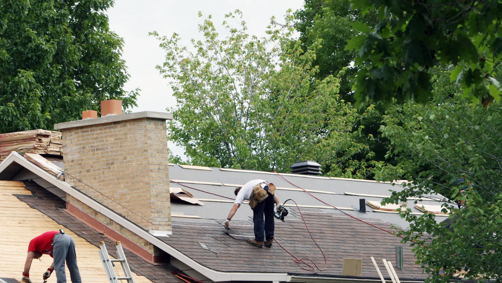 Two roofers installing shingles on a house beside a brick chimney